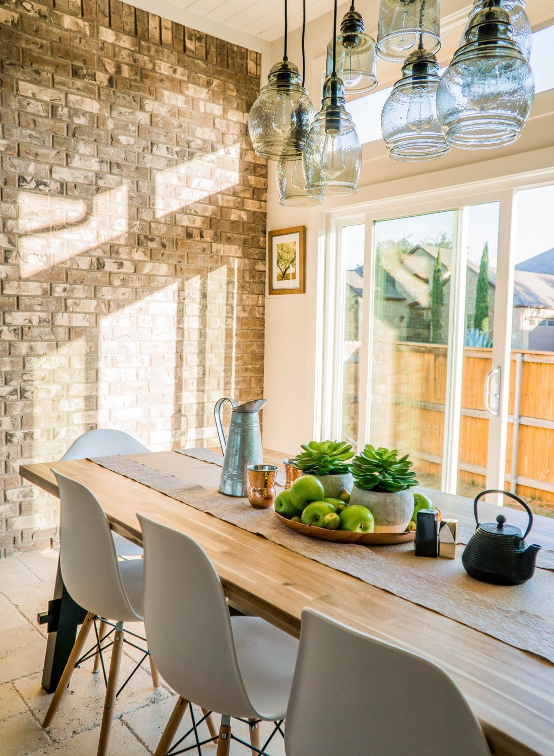 Long farmhouse table with mid-century modern white chairs against brick wall and oversized windows